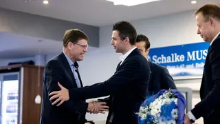 Grütters und Kirstein begrüßen sich per Handschlag bei einer Veranstaltung; im Vordergrund ein Strauß mit blauen und weißen Blumen, im Hintergrund ein Schild zum Schalke‑Museum.