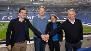Der Vorstand der Auf Schalke eG im Schalke‑Stadion legen die Hände übereinander; im Hintergrund das Spielfeld und die Tribüne mit dem Schriftzug „FC SCHALKE 04“